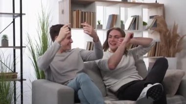 Expressive joy. Funny men. Friendly relationship. Happy casual handsome guys having fun dancing with hands up sitting sofa in light room interior slow motion.