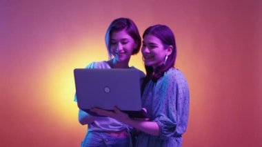 Digital chatting. Flirting women. Neon light people. Happy curious female friends typing laptop posing purple orange background.