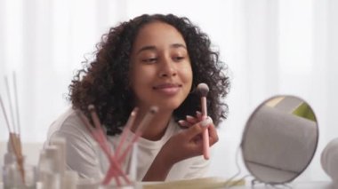 Morning makeup. Beauty care. Decorative cosmetics. Smiling pretty girl applying blush with brush on radiant face skin looking in mirror.