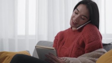 Mobile connection. Shopping list. Female housekeeper. Smiling woman making notes in copybook talking smartphone sitting sofa in light room interior.