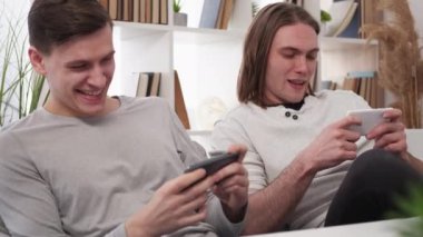 Interesting game. Competitive men. Home leisure. Happy male friends playing together by smartphones sitting sofa in light room interior.