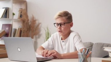 Home education. Smart boy. Online conference. Inspired teenager kid in spectacles talking laptop sitting desk light room interior.