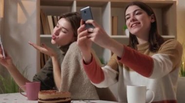 Selfie life. Coquettish women. Home meeting. Happy female friends making photo on smartphone flirting and sending kisses sitting light room interior.