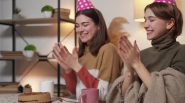 Birthday greeting. Happy women. Meeting joy. Cheerful female friends in festive hat clapping hands sitting desk with sweet cake in light room interior.