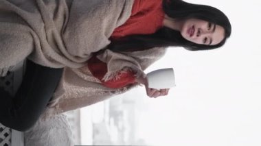 Dikey video. Sabah kahvesi. Rahat kadın. Keyifli bir an. Mutlu kadın pencere pervazında oturmuş, ekoseyi iç mekânla kaplayan içecek içiyor..