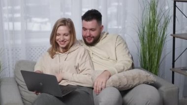 Online call. Happy couple. Distance meeting. Smiling relaxed man and woman talking to laptop sitting together on sofa light room interior.