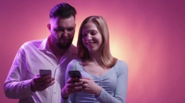 Mobile game. Funky couple. Online entertainment. Happy woman and man playing on personal smartphones having fun together posing neon light orange purple background.