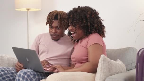 假期快乐 一对快乐的夫妇网上旅行 兴奋的黑人男人和女人坐在明亮房间的沙发上 获得意想不到的令人愉快的信息 — 图库视频影像