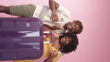 Vertical video. Happy vacation. Beloved couple. Travelling joy. Joyful black man and woman showing thumbs up posing suitcase and cocktails on pink background.