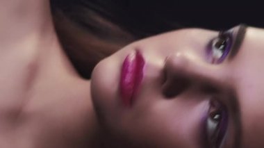 Vertical video. 90s beauty. Retro makeup. 2000s fashion. Closeup portrait of teen girl face with purple color eyeshadow pink lips artistic look posing in neon light on dark black.