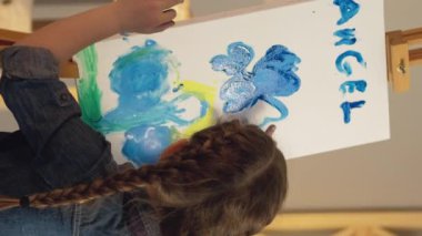 Vertical video. Child art. Creative hobby. Talent inspiration. Little girl finger painting blue yellow color angel artwork on easel.