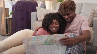 Vacation planning. Excited couple. Romantic trip. Happy inspired black man and woman studying map sitting floor with packed suitcase light room interior.