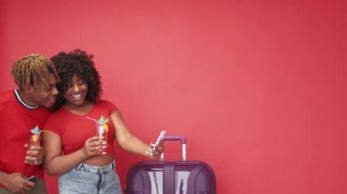 Cep telefonu fotoğrafı. Romantik tatil. Heyecanlı bir çift. Mutlu siyah adam ve kadın, kırmızı arka planda duran akıllı telefondan selfie bavulu ve kokteyl yapıyorlar..