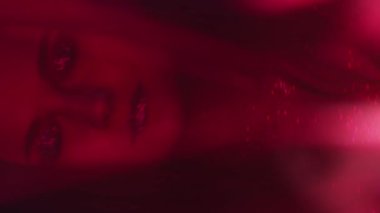 Vertical video. Color light face. Nightclub beauty. Fashion look. Blur neon red woman with shimmering glitter artistic makeup metallic eyeshadow lips dancing on dark.