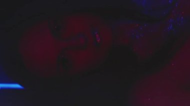 Vertical video. Neon girl. Nightclub makeup. Glamour look. Red blue color light woman with sparkling glitter face skin metallic eyeshadow lips dancing on dark.