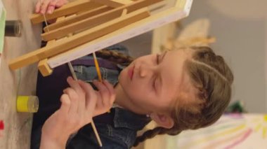 Vertical video. Art school. Kid talent. Creative leisure. Female teacher little girl painting together with paintbrushes on easel in studio.