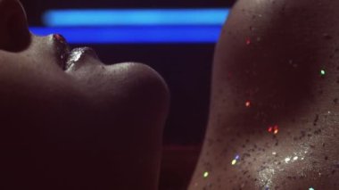 Vertical video. Glitter skin. Makeup art. Nightclub look. Cropped closeup of neon blue red color light woman face with sparkling metallic silver lips on dark.
