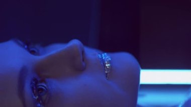 Vertical video. Glitter makeup. Neon girl. Nightclub fashion. Blue purple color flash light woman face with sparkling metallic eyeshadow lips in mirror on dark.