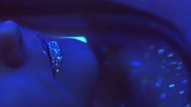 Vertical video. Disco makeup. Neon girl. Nightclub beauty. Cropped closeup of blue color light woman face with sparkling glitter skin metallic lips on dark.