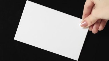 Vertical video. White card. Empty note. Invitation letter. Female hand showing clean mockup paper sheet with copy space on black background loop.
