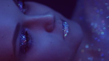 Vertical video. Neon face. Glitter makeup. Glamour beauty. Closeup portrait of purple color light woman with sparkling skin eyeshadow lips on dark.