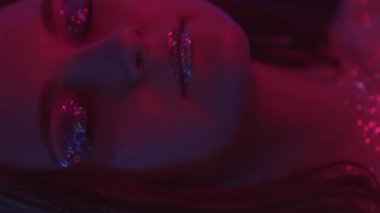 Vertical video. Neon face. Glitter makeup. Disco fashion. Red color light relaxed woman with sparkling skin metallic eyeshadow lips enjoying dancing on dark.