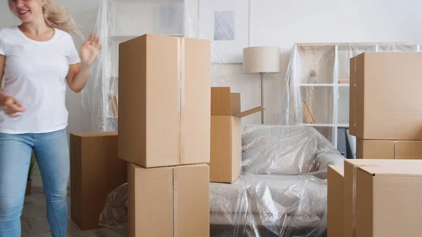 Home moving. Happy relocation. Package delivery. Defocused smiling woman arranging boxes in light flat with polyethylene covered furniture.