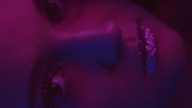 Vertical video. Neon face. Sparkling makeup. Disco vogue. Closeup of pink blue color light girl with shiny glitter skin metallic eyeshadow lips on dark.