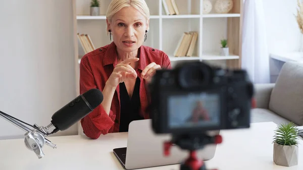 Video blog. Interview streaming. Online expert. Senior influencer woman speaking into microphone recording vlog on camera on tripod at modern home interior.