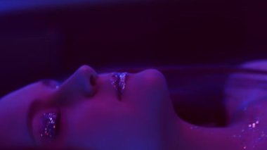 Vertical video. Neon girl. Glitter makeup. Nightclub beauty. Purple color light relaxed woman with sparkling face skin eyeshadow lips dancing with closed eyes on dark.