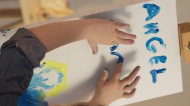 Vertical video. Finger painting. Kid art. Little artist imagination. Talented girl hands creating blue yellow color angel heart artwork on easel.