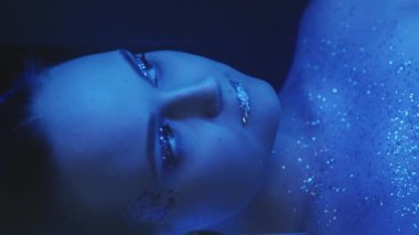 Vertical video. Sparkling makeup. Neon beauty. Disco look. Night portrait of blue color light woman face with shiny glitter skin eyeshadow lips on dark.