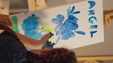 Vertical video. Kid creativity. Painting art. Young artist talent. Inspired small girl blowing off glitter from hands on blue yellow color angel artwork.