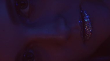 Vertical video. Night makeup. Neon face. Disco look. Closeup of pink blue color light glitter girl with sparkling skin metallic eyeshadow lips on dark.