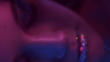 Vertical video. Neon beauty. Party makeup. Nightclub look. Closeup portrait of purple blue color light woman face with shimmering eyeshadow lips on dark.