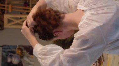 Vertical video. Artist lifestyle. Art therapy. Creative hobby. Inspired woman fixing hair with paintbrushes painting abstract picture on canvas at home studio.