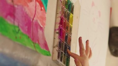 Vertical video. Finger painting. Kid art. Young artist. Child hand mixing colors in palette sprinkling watercolor paint over abstract artwork.