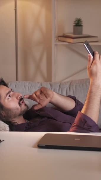 垂直视频呼叫 在线聊天 远程通信 躺在现代家庭内部沙发上用手机摄像头说话的松懈男人 — 图库视频影像
