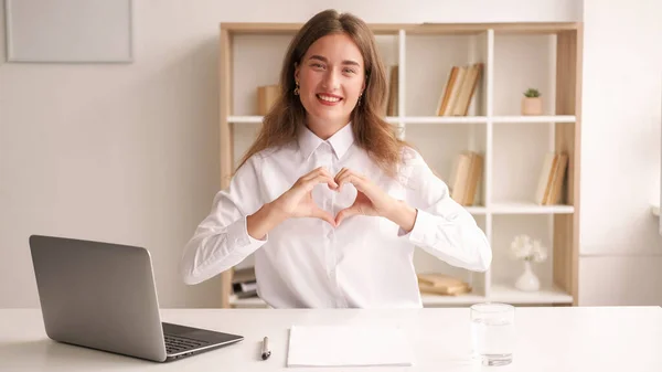 Sending love. Office woman. Inspired work. Smiling lady in white shirt sitting desk with laptop showing heart shape figure hands in light room interior.