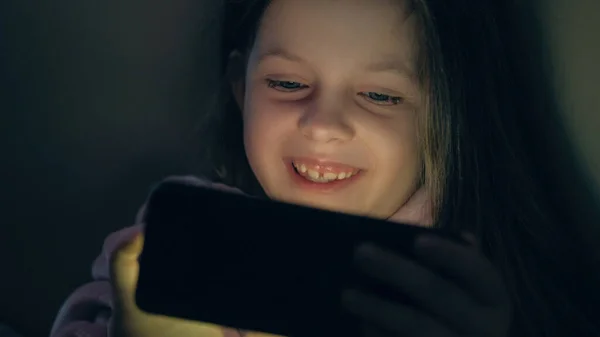 Child night movie. Gadget kid. Late fun. Entertained happy smiling little girl enjoying watching video cartoons on phone laughing in dark.