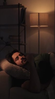 Dikey video. Huzursuz bir gece. Uykusuzluk sorunu. Zihinsel aşırı yüklenme. Üzgün, düşünceli, düşünceli bir adam geç saatlere kadar kanepede uzanıyor..