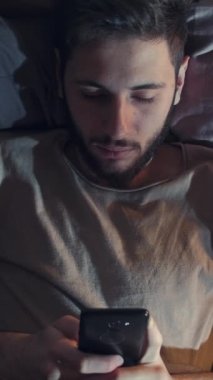 Dikey video. Gadget gecesi. Mobil sohbet. İnternet uykusuzluğu. Sıkılmış yorgun adam mesajlaşan mesajlar kullanıyor. Karanlık yatak odasında geç saatte yatakta yayını kaydırıyor..