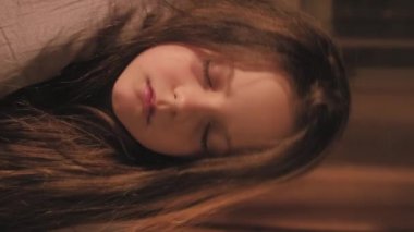 Dikey video. Yorgun çocuk istirahati. Gece yorgunluğu. Yorgun, gözleri battaniyeli küçük bir kız karanlık bir yatak odasında rahat bir yatakta uyuyakalıyor..