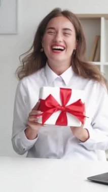 Dikey video. Şirket selamlaması. Heyecanlı kadın. Beklenmedik bir sürpriz. Mutluluğunu ifade eden mutlu bir bayan elinde hediye kutusu kırmızı kurdele oturma masasında iç mekânda oturuyor..