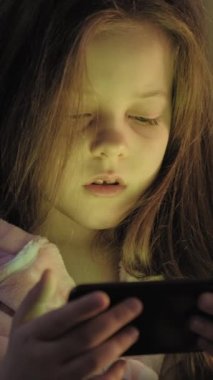 Dikey video. Uyku zamanı aleti. Çocuk uykusuzluğu. Yorgun küçük kız çizgi film seyrediyor gecenin geç saatlerinde yatakta telefon kullanıyor..