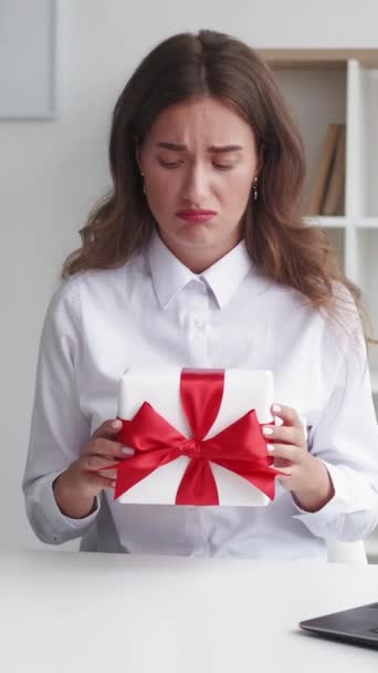 垂直录像 坏礼物 让人失望的女人企业问候 心烦意乱的女士不满意地拿着礼物盒 红丝带 坐在明亮的房间里 — 图库视频影像
