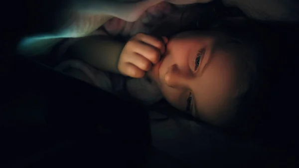 Kid bed movie. Night gadget. Bedtime entertainment. Curious impressed small girl enjoying watching video cartoons under blanket late in dark.