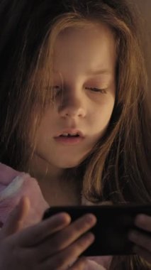 Dikey video. Uyku zamanı aleti. Çocuk uykusuzluğu. Yorgun küçük kız çizgi film seyrediyor gecenin geç saatlerinde yatakta telefon kullanıyor..