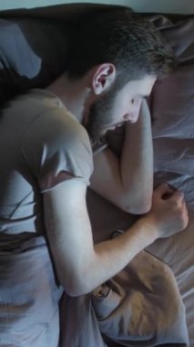 Dikey video. Uyku sorunu. Gece uykusuzluğu. Dinlenme problemi. Endişeli bir adam, karanlık bir yatak odasında yalnız başına yatakta dönüp duruyor..