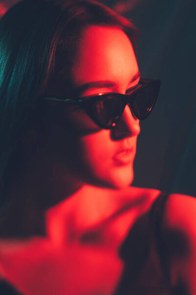 Eyewear fashion. Art portrait. Optical accessories. Red color light defocused woman face wearing black stylish sunglasses on dark.